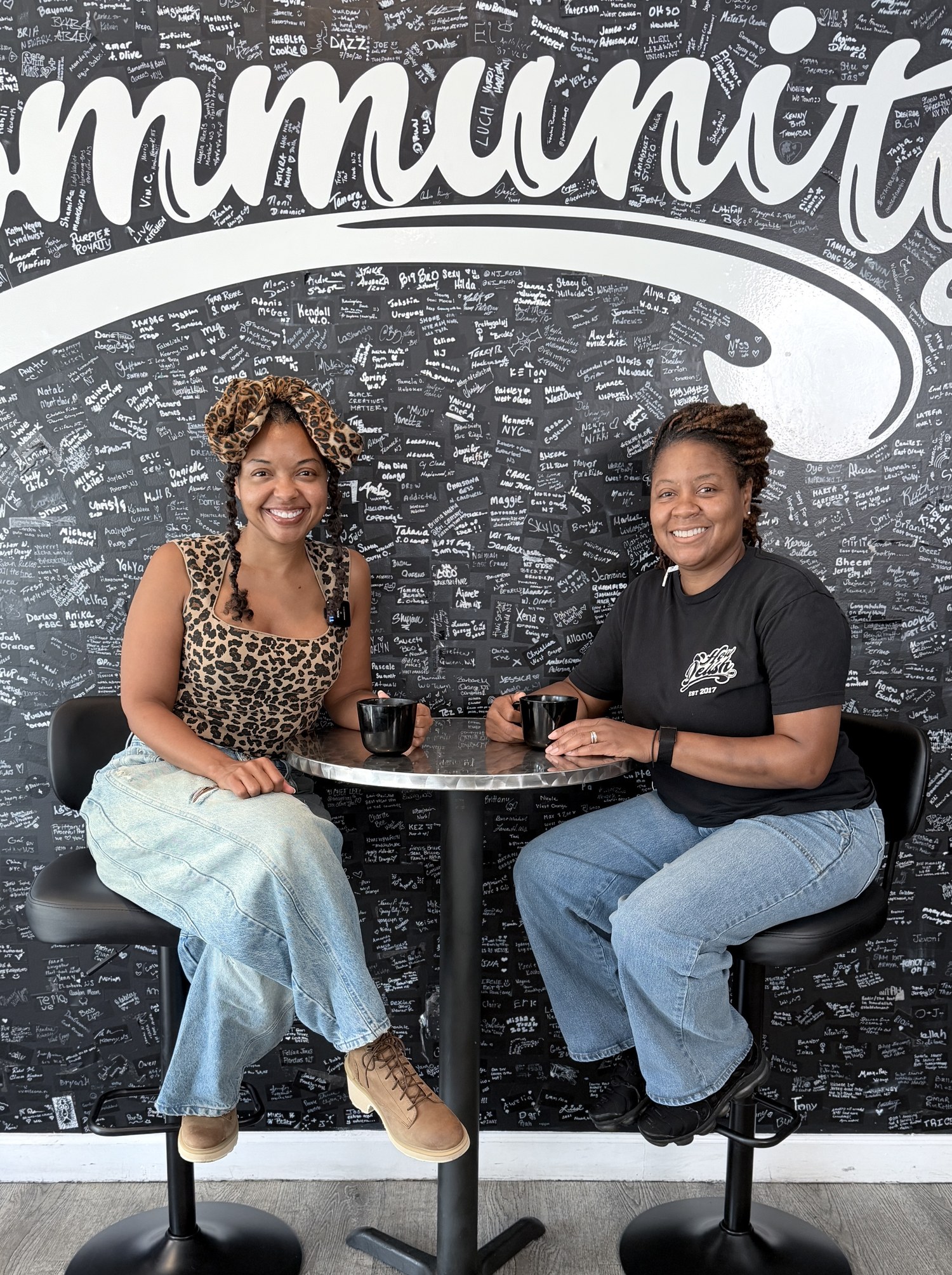 Two women seated at a cafe table in front of a Community mural wall